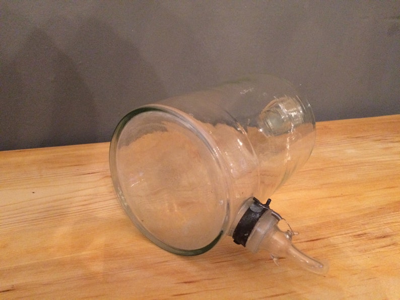 May include: A clear glass bottle with a spout and a black plastic cap. The bottle is lying on its side on a wooden surface.