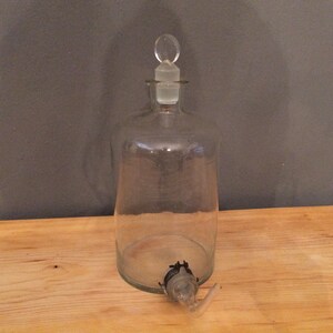 May include: A clear glass bottle with a glass stopper and a spout. The bottle is empty and has a textured surface.