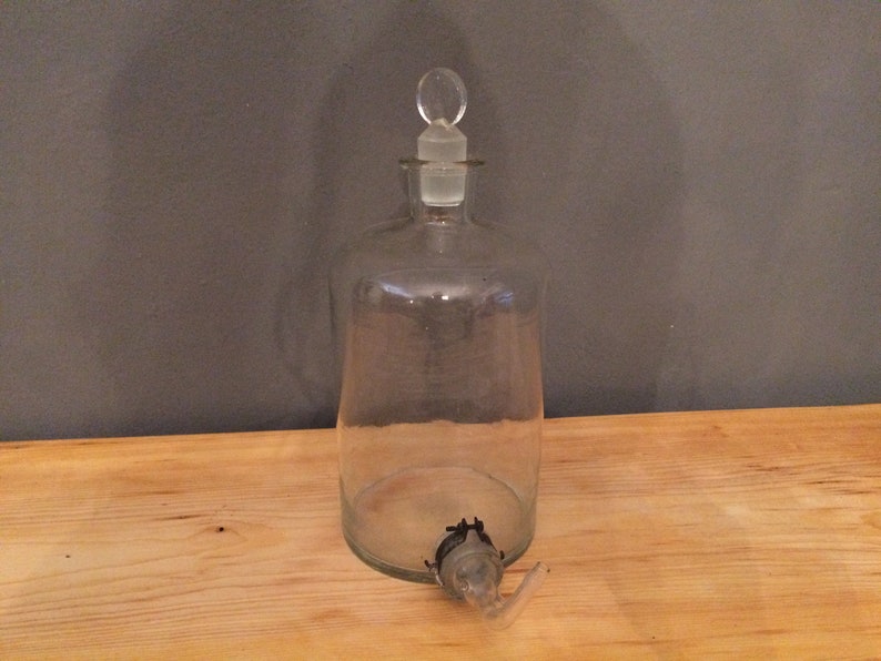 May include: A clear glass bottle with a glass stopper and a spout. The bottle is empty and has a textured surface.