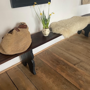 Könnte beinhalten: Rustikale Holzbank mit dunkelbrauner Beize und schwarzen Beinen. Eine große gewebte Tragetasche mit Lederakzenten liegt auf der Bank. Eine Vase mit gelben Narzissen und ein Schaffell vervollständigen die Szene.