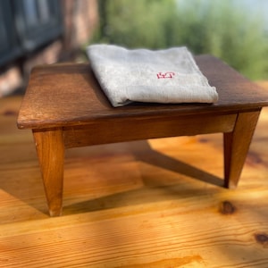 Könnte beinhalten: Ein kleiner, rechteckiger Holzhocker mit vier konisch zulaufenden Beinen und einer glatten, mittelbraunen Oberfläche. Auf dem Hocker liegt ein gefaltetes, hellbeiges Leinentuch mit einem kleinen, roten Monogramm der Buchstaben LP.