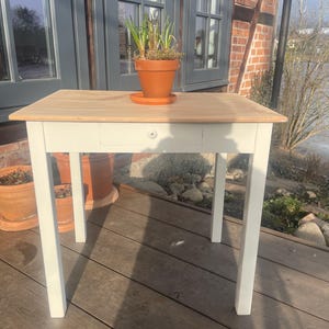 Könnte beinhalten: Kleiner Holztisch mit hellbrauner Tischplatte und weißen Beinen. Auf der Tischplatte steht ein Terrakotta-Topf mit einer Pflanze. Der Tisch hat eine Schublade. Er steht im Freien auf einer Holzterrasse.