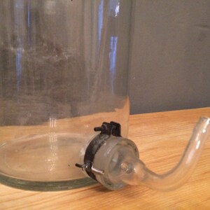 May include: A clear glass bottle with a glass spout and a metal clamp. The bottle is sitting on a wooden surface.