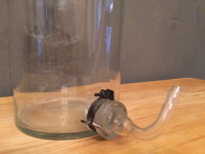 May include: A clear glass bottle with a glass spout and a metal clamp. The bottle is sitting on a wooden surface.