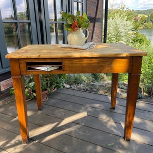 Op de afbeelding: Een rechthoekige houten tafel met een natuurlijke lichtbruine afwerking, voorzien van vier rechte poten en een open opbergvak in de voorste rand. Bovenop staat een witte keramische kan gevuld met groen blad en rode bessen.