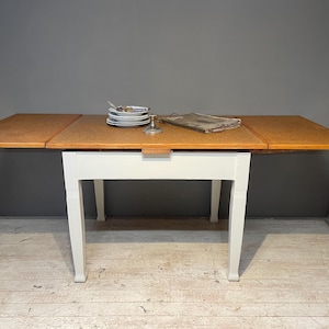 Könnte beinhalten: Ein Esstisch aus Holz mit hellbrauner Tischplatte und weißen Beinen. Der Tisch hat zwei ausgeklappte Klappen. Ein Stapel weißer Teller, ein kleiner silberner Kerzenhalter und eine Zeitung liegen auf dem Tisch.