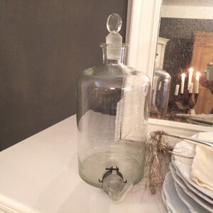 May include: A clear glass bottle with a glass stopper and a spout. The bottle is sitting on a white surface.