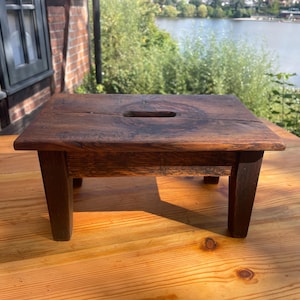 Könnte beinhalten: Ein kleiner, rechteckiger Holzhocker mit einer dunkelbraunen, verwitterten Oberfläche. Die Sitzfläche verfügt über einen markanten, mittig platzierten horizontalen Griffschlitz. Der Hocker steht auf vier stabilen, geraden Holzbeinen.