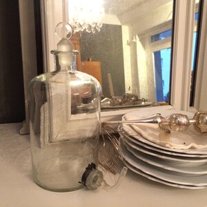 May include: A clear glass bottle with a stopper and a silver and white decorative ornament on a white surface.