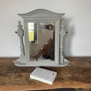 Peut inclure: Un miroir de toilette en bois gris avec un miroir rectangulaire et deux colonnes décoratives de chaque côté. Le miroir est posé sur une surface en bois.