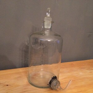 May include: A clear glass bottle with a glass stopper and a glass spout. The bottle is empty and has a textured surface.
