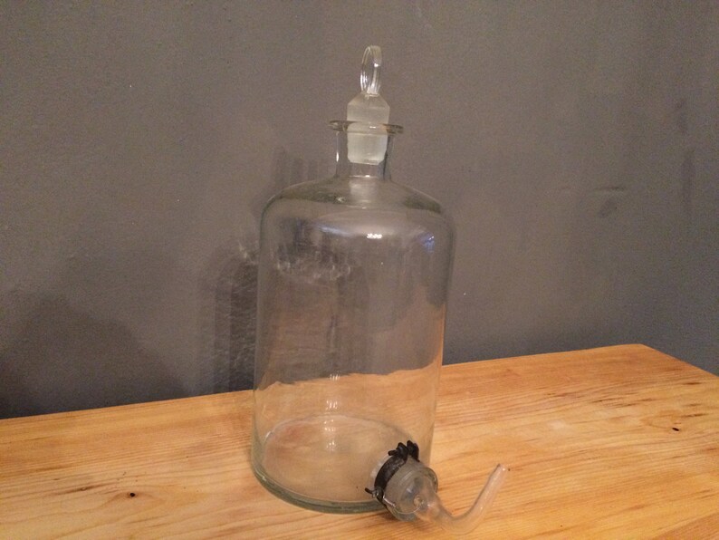 May include: A clear glass bottle with a glass stopper and a glass spout. The bottle is empty and has a textured surface.