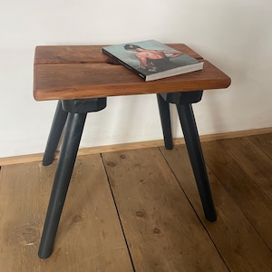 Peut inclure: Un petit tabouret en bois avec un dessus rectangulaire et trois pieds noirs. Le dessus est de couleur brun chaud, et un livre avec une image en couleur repose dessus. Le tabouret est sur un plancher en bois.