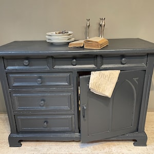 Könnte beinhalten: Ein dunkelgrauer Holzschrank mit Schubladen und einer offenen Tür. Der Schrank hat eine rechteckige Oberseite mit einer leicht überstehenden Kante. Obenauf befinden sich weiße Teller, Bücher und Kerzenständer. Der Schrank hat eine Vintage-Ästhetik.