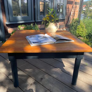 Op de afbeelding: Een houten tafel met een donker gebeitst blad en zwarte poten. Een open boek en een crèmekleurige kan met bloemen staan op de tafel. De tafel staat op een houten terras.