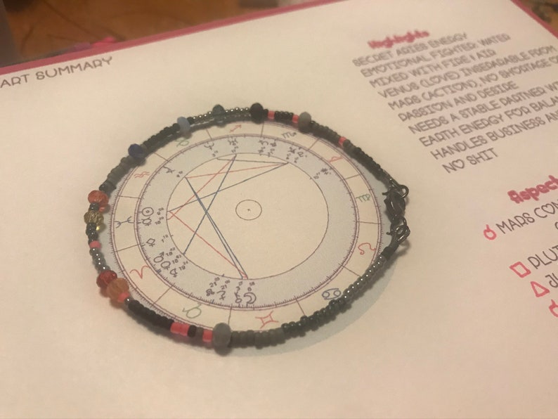 K&ouml;nnte beinhalten: Ein schwarzes Perlenarmband mit bunten Perlen, darunter rote, rosa, blaue und goldene. Das Armband liegt auf einem wei&szlig;en Papier mit einem schwarz-wei&szlig;en astrologischen Diagramm darauf gedruckt. Der Text auf dem Papier lautet "Highlights", "Secret Aries Energy", "Emotional Fighter Water", "Mixed with Fire I Aid", "Venus (Love) Ingedable from", "Mad's Caction), No Shortageace", "Passion and Decide", "Needs a Stable Dartner Wo", "Earth Energy for Balan", "Handles Business An", "No Shit", "Aspect", "Mars Con", "Plu", "&Delta;&alpha;".