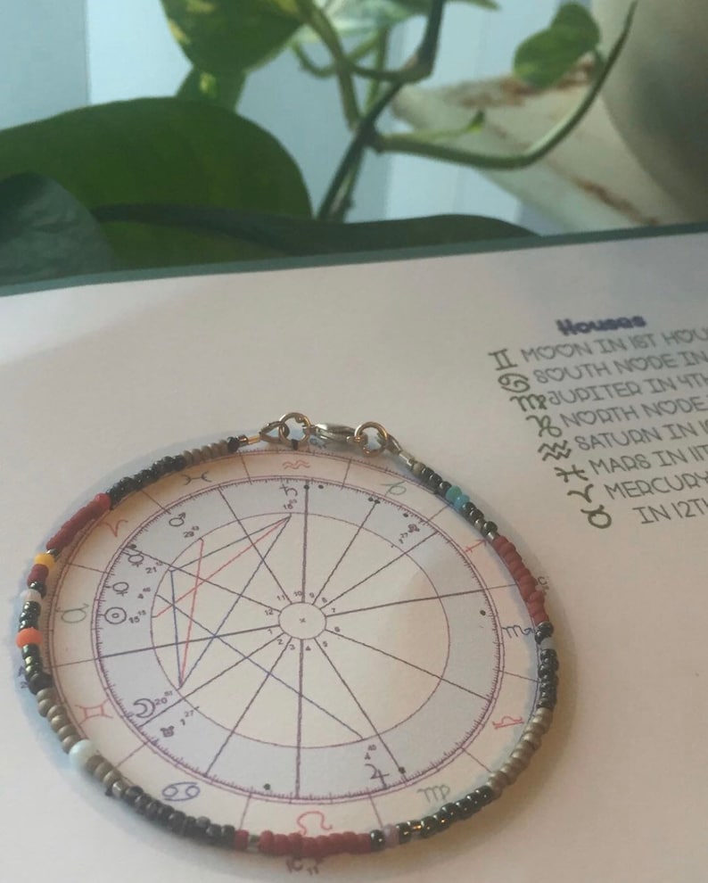 K&ouml;nnte beinhalten: Ein Armband aus Perlen in verschiedenen Farben, darunter Rot, Orange, Gelb, Blau und Grau. Das Armband liegt auf einem Papier mit einem Horoskop und Text, der "H&auml;user II Mond im 1. Haus S&uuml;dlicher Knoten in MJupiter im 4. Haus Nordischer Knoten Saturn in IS Mars in IIT Merkur in 12T" lautet.