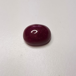 Può includere: Una gemma lucida, di forma ovale, di colore rosso intenso. La gemma ha una superficie liscia e riflettente, con una sottile lucentezza. Il colore è un rosso scuro intenso, con sfumature di viola. La gemma è su uno sfondo bianco.
