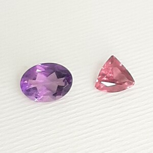 May include: Two gemstones: an oval-cut amethyst and a triangular pink tourmaline. The amethyst is a deep purple with internal facets. The tourmaline is a lighter pink with a triangular shape and a faceted surface. Both are set against a white background.