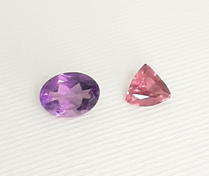 May include: Two gemstones: an oval-cut amethyst and a triangular pink tourmaline. The amethyst is a deep purple with internal facets. The tourmaline is a lighter pink with a triangular shape and a faceted surface. Both are set against a white background.
