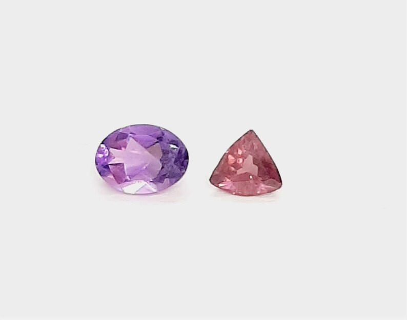 May include: An oval-cut amethyst gemstone and a triangular pink tourmaline gemstone are displayed against a white background. The amethyst is a deep purple color, while the tourmaline is a vibrant pink.
