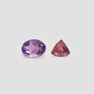 May include: Two gemstones on a white background. One is an oval-cut amethyst in shades of purple. The other is a triangular, pink gemstone. Both stones are faceted and reflect light.