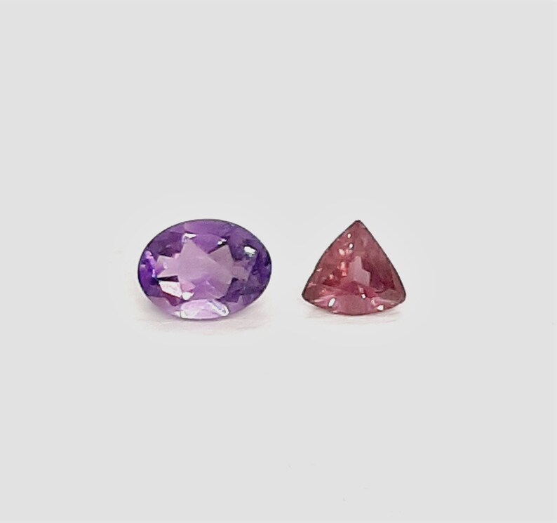 May include: Two gemstones on a white background. One is an oval-cut amethyst in shades of purple. The other is a triangular, pink gemstone. Both stones are faceted and reflect light.
