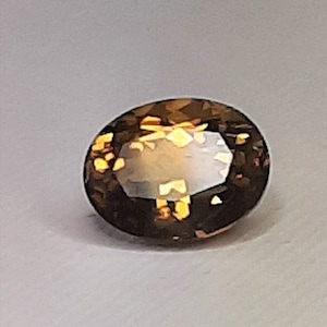 Puede incluir: Una gema de corte ovalado, de color marrón oscuro, con destellos dorados internos. La gema está facetada, reflejando la luz y mostrando su claridad. Adecuada para joyería.