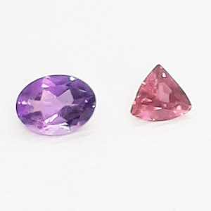 May include: Two gemstones on a white background. One is an oval-cut amethyst in shades of purple. The other is a triangular-cut pink gemstone. Both stones are faceted, reflecting light.