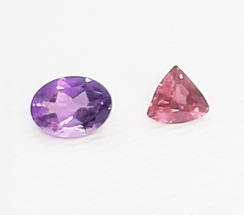 May include: Two gemstones on a white background. One is an oval-cut amethyst in shades of purple. The other is a triangular-cut pink gemstone. Both stones are faceted, reflecting light.