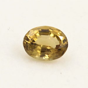 Puede incluir: Una gema ovalada de color amarillo dorado. La gema tiene una superficie facetada que refleja la luz, creando un efecto brillante. La piedra se encuentra sobre un fondo blanco liso, resaltando su color y claridad. Esta gema podría usarse en joyería.