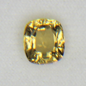 Puede incluir: Una gema de color amarillo dorado con talla cojín. La gema tiene una superficie facetada que refleja la luz, creando un efecto brillante. La piedra está sobre un fondo blanco.