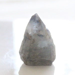 Puede incluir: Una punta de cristal gris con una punta ligeramente translúcida. La piedra tiene una textura rugosa y natural y un sutil degradado de color, que pasa de una base más oscura a una parte superior más clara. El cristal se muestra sobre un fondo blanco.