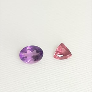 May include: Two gemstones on a white surface. One is an oval-cut amethyst, displaying shades of purple. The other is a triangular, pink gemstone. Both stones are faceted, reflecting light.