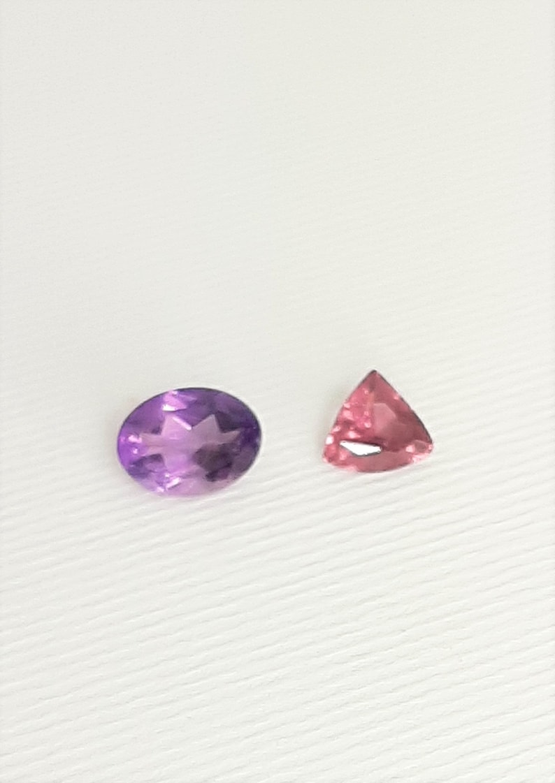 May include: Two gemstones on a white surface. One is an oval-cut amethyst, displaying shades of purple. The other is a triangular, pink gemstone. Both stones are faceted, reflecting light.
