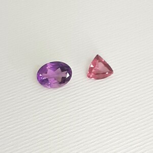 May include: Two gemstones on a white surface. One is an oval-cut amethyst with a deep purple hue. The other is a triangular-cut pink gemstone. The gemstones are displayed against a plain white background.