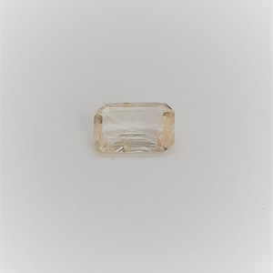 May include: A rectangular, light-colored gemstone with a subtle, warm hue. The gem has a clear, transparent appearance with visible internal facets. The cut is a classic emerald shape, showcasing its clarity and brilliance. The stone is set against a white background.