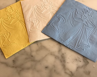 May include: Three paper napkins with an embossed image of Cinderella in a ball gown. The napkins are yellow, white, and light blue.