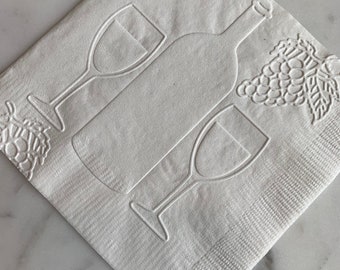 Puede incluir: Servilleta de papel blanca con un diseño en relieve de una botella de vino, dos copas de vino y un racimo de uvas.