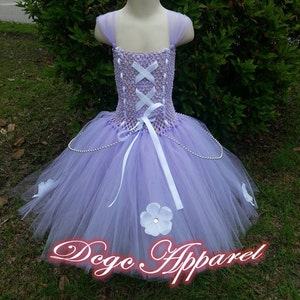 May include: A lavender tutu dress with a lace-up bodice and tulle skirt. The dress features white ribbon accents, a white flower, and a pearl trim. The text "Dego Apparel" is displayed in red cursive.