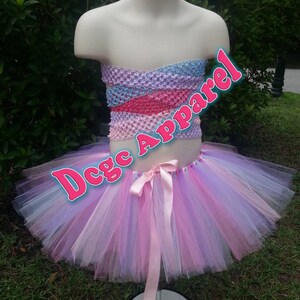 May include: A multi-colored tutu set on a mannequin. The top is a combination of blue, pink, and purple. The tutu skirt is layered with pink, purple, and white tulle. A pink ribbon is tied at the waist. Text on the image reads "Dcgc Apparel".
