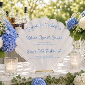 Puede incluir: Un letrero blanco en forma de concha con texto azul que enumera cócteles exclusivos: Katie's Aperol Spritz y Eric's Old Fashioned. El letrero está apoyado en un soporte dorado, con arreglos florales y velas en el fondo.