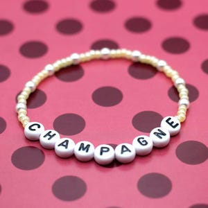 May include: A beaded bracelet with the word "CHAMPAGNE" spelled out in white letter beads. The bracelet has a cream-colored band with silver beads and is set against a pink background with dark red polka dots.