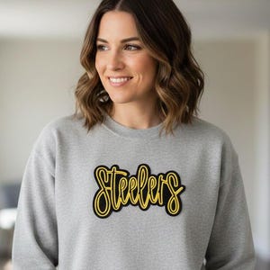 May include: A gray sweatshirt with the word "Steelers" in yellow script with a black outline. The sweatshirt is a crew neck style.