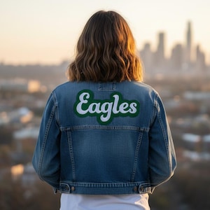 May include: A denim jacket with the word "Eagles" in white script with a green outline on the back. The jacket is blue and has a classic design. The background shows a city skyline at sunset.