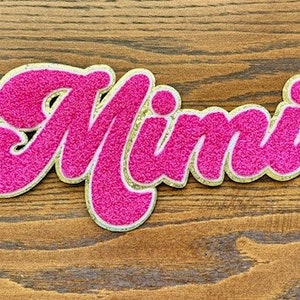 May include: Pink glitter iron-on patch with the word "Mimi" in a cursive font with a gold outline.