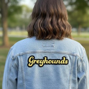 May include: Light blue denim jacket with the word "Greyhounds" in yellow and black lettering on the back. The jacket is worn by a person with brown hair, with a blurred green background.