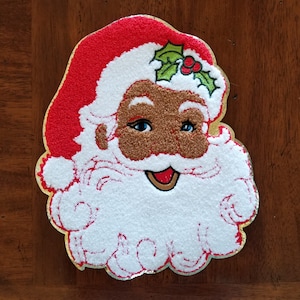 May include: A festive Santa Claus face patch with a red hat and gold trim. The face features a brown complexion, blue eyes, and a white beard. Holly and berries adorn the hat. The patch is likely for holiday crafts.