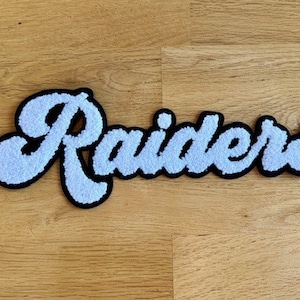 May include: White fluffy patch with the word "Raiders" in black outlined lettering.
