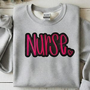 May include: A light gray sweatshirt with the word "Nurse" in pink script with a black outline. A small pink heart is next to the word. The sweatshirt has a crew neck and long sleeves.
